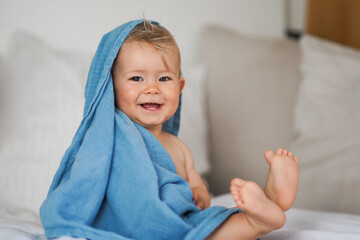 mega cute naked almost one year old blond baby boy sitting & laughing at home on a cozy bed after bathing and playing with a blue muslin fabric burp cloth while making nonsense jokes - obrazy, fototapety, plakaty