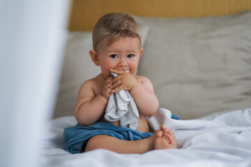 cute naked almost one year old blond baby boy sitting at home on a cozy bed after bathing and...