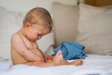 cute naked one year old happy blond baby boy sitting at home on a cozy bed after bathing and looking down to his genitals and discovering his body