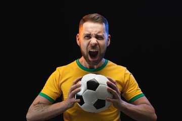 Studio image of excited screaming supporter man in yellow t-shirt cheering for his favourite team, posing over black background with a soccer ball in hands