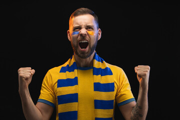 Portrait of euphoric happy soccer supporter man in yellow-blue t-shirt and scarf and painted face, cheering for his favourite team, isolated over black background - 527436498