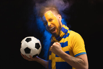 Portrait of excited soccer supporter with yellow-blue painted face is posing with a ball in hands over black background with smoke effect