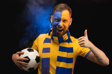 Portrait of happy excited soccer fan with yellow-blue painted face is holding ball in hand and showing thumbs up gesture, while posing over black background with smoke effect