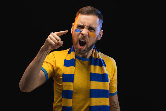 Portrait of excited young soccer supportred man in yellow-blue t-shirt and scarf and painted face, cheering for his favourite team, isolated over black background