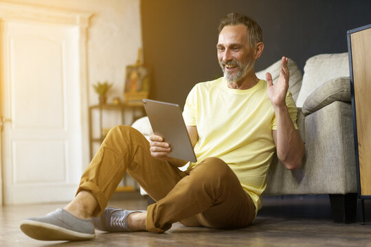 Happy grey haired man has video chat on tab computer in hand while sitting in living foom floor near of sofa. Handsome bearded mature man on floor indoor gesture by hand hi via internet communication