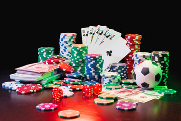 Studio shot composition of poker chips, dices, playing cards, soccer ball and dollar banknotes on the desk with grunge texture over black background. Casino, poker, gambling, sports betting, money win