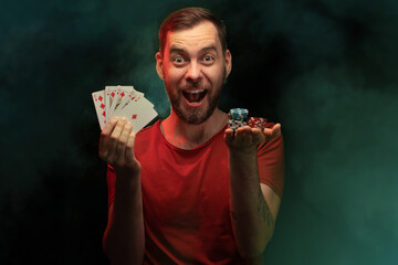 Studio shot of happy smiling man posing with a stack of playing cards and a pile of poker chips in...