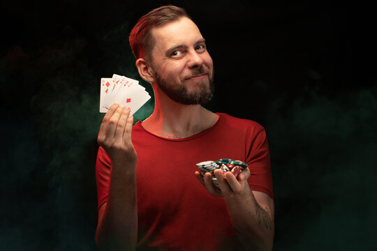 Studio portrait of smiling handsome young bearded man posing with a fan of playing cards and pile of poker chips over black background with smoke effect