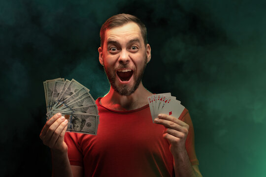 Studio Portrait Of Young Euphoric Man Posing With A Stack Of Playing Cards And Ward Of Dollar Banknotes In Hands Over Black Background With Smoke Effect