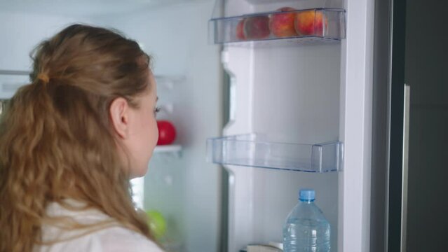 Woman putting groceries in new modern fridge. Young adult female unpacking shopping bag loading modern hi-tech refrigerator. Girl putting fruit and vegetables into fridge closeup shot