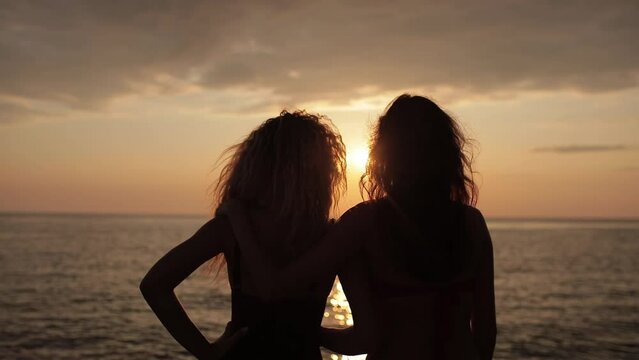 Silhouette of couple friends at sea in golden and beautiful sunset. Girls on holidays on romantic summer evening. Happy couple talking and hugging on beach