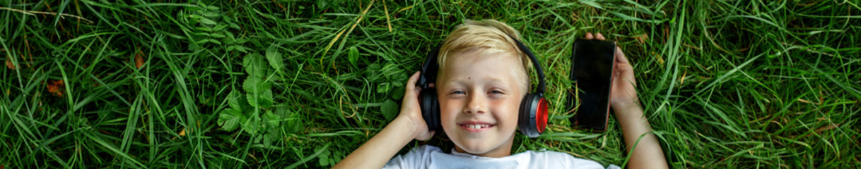 Child listens to music. Blond boy uses headphones and smart phone, lies in grass. Top view. Banner