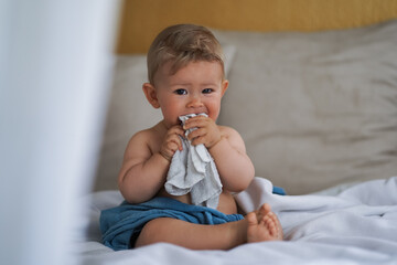 cute naked almost one year old blond baby boy sitting at home on a cozy bed after bathing and playing or biting a white flannel face cloth because of teething and suck water