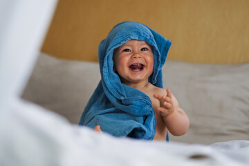 Naklejka premium mega cute naked almost one year old blond baby boy sitting & laughing at home on a cozy bed after bathing and playing with a blue muslin fabric burp cloth while making nonsense jokes