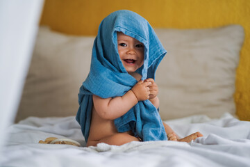 mega cute naked almost one year old blond baby boy sitting & laughing at home on a cozy bed after bathing and playing with a blue muslin fabric burp cloth while making nonsense jokes - obrazy, fototapety, plakaty