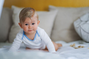 Fototapeta premium cute happy one year old happy blond babyboy in a white body or suit at home on a cozy bed after bathing and looking into the Camera 