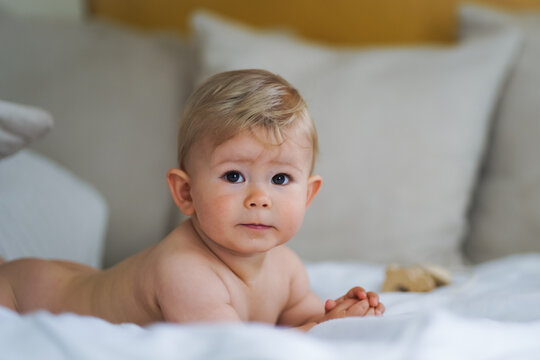 super sweet naked almost one year old happy blond baby boy lying at home on a cozy bed after bathing in prone position and smiling or flirting with the Camera like a innocent rascal