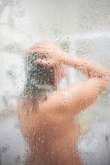Blurred middle-aged unrecognizable caucasian woman enjoying hot shower in steamy bathroom behind the focused water splashed glass of the shower cabin.