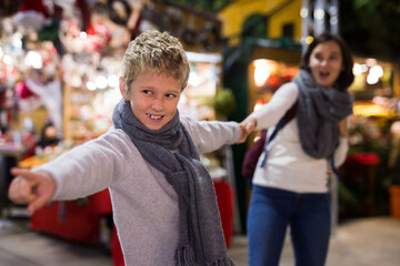 Obraz na płótnie Canvas Preteen boy pulling his mom hand, demanding to buy something for him at outdoor Christmas market