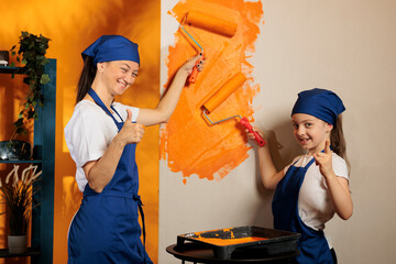 Mother and girl painting and giving thumbs up, using orange color paint and roller bursh to redecorate apartment. People doing okay like approval gesture and renovating room walls.