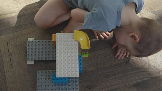 caucasian child boy kid building house with colorful plastic bricks parts. preschooler sitting on wooden floor playing with blocks constructor. child developing imagination, architecture skills, build