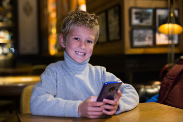 Smiling preteen boy sitting at table in cozy empty cafe, playing with smartphone