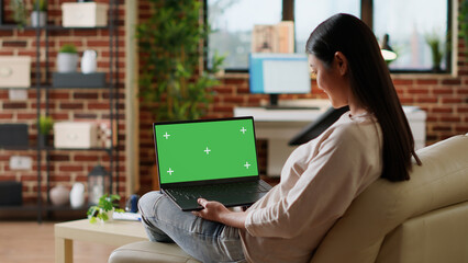 Young woman sitting at home with laptop having green screen chroma key display. Adult person sitting on sofa with portable computer on lap having mockup isolated template background. Tripod shot - Powered by Adobe