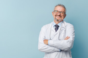 Smiling male doctor in lab coat with arms crossed against blue background.