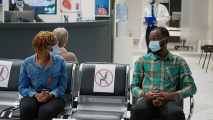 African american patients with face mask talking in waiting area, preparimg to start checkup examination with medic during covid 19 pandemic. Man and woman sitting in hospital reception.