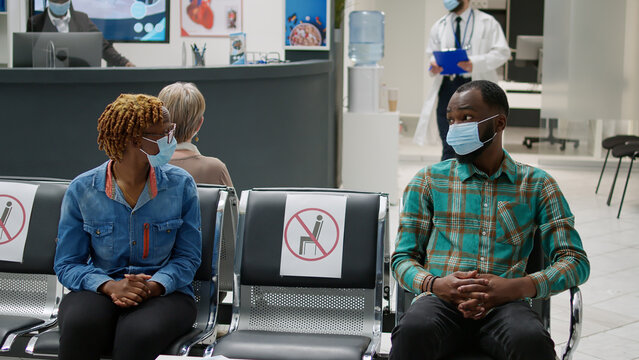 African american patients with face mask talking in waiting area, preparimg to start checkup examination with medic during covid 19 pandemic. Man and woman sitting in hospital reception. - Powered by Adobe