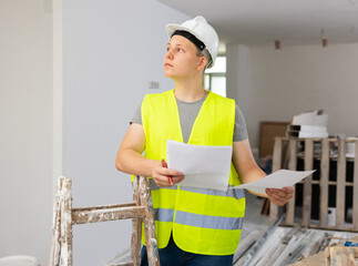 Teenage boy in yellow vest and hardhat checking documents in construction site during workday on...