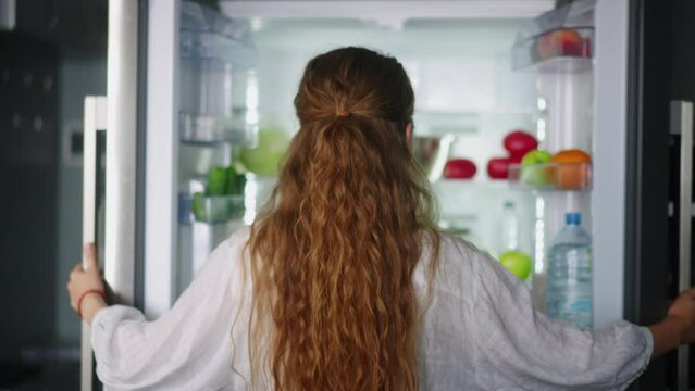 Young woman taking vegetables from fridge and has fun singing and dancing in modern minimalistic kitchen with island. Girl getting tomato from side by side refrigerator and singing to cucumber.