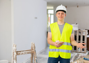 Portrait of teenage boy wearing yellow vest and hardhat holding bunch of earned money for part-time work in construction site.