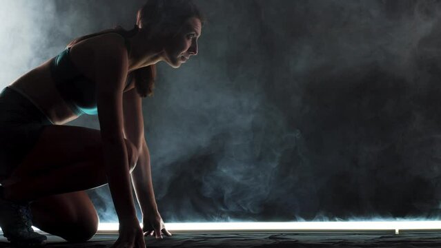 Silhouette female runner in race  start position. Girl in sportswear posing on lit track with smoke in the background.