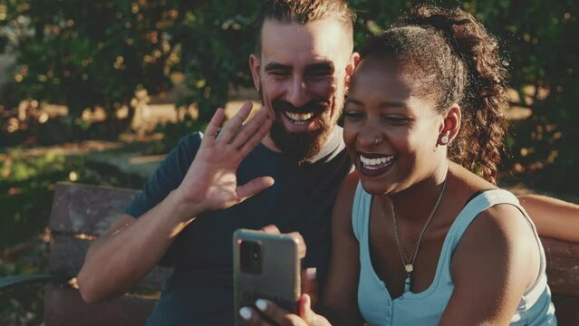 Happy smiling interracial couple making video call on cellphone while sitting on bench