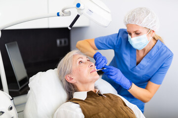 Experienced female cosmetologist working with senior female client in clinic of aesthetic medicine, injecting facial filler into skin to correct face contour