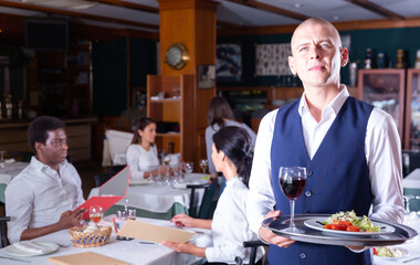 Hospitable waiter standing with serving tray in cozy restaurant hall, welcoming guests