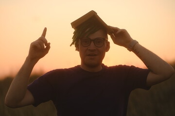 Young puzzled man in glasses with dreadlocks holding book above head as roof, raises his index...
