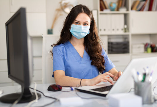 Young Trainee Doctor In A Protective Mask, Undergoing An Internship At A Clinic During A Pandemic, Sits At A Desktop ..in Front Of A Computer In Office