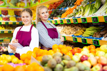 Portrait of confident smiling saleswoman standing with trainee salesgirl among shelves with fresh organic fruits and vegetables in grocery store
