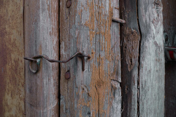 An old brass hook closes a dilapidated door on an old dilapidated wooden shed. The paint is peeling off the boards and rusty nails are sticking out.