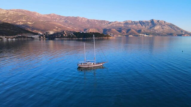 Scenic top view sailing small yacht sailing in turquoise Adriatic Sea. Aerial drone tracking video. Ship is sailing on sea. Montenegro mountains, sea, island. Image of ideal seaside vacation, cruise.