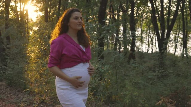 Pregnant mom looking ahead while walking in forest as exercise, sun sets behind