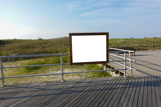 Blank rectangular wooden frame with white center attached to railing on a wooden boardwalk by a beach with a sand dune and grass in the background