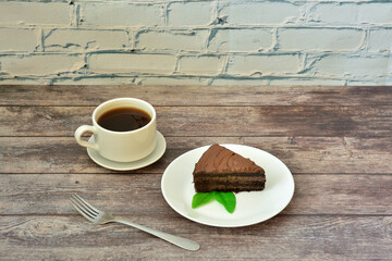 A plate with a piece of chocolate cake with mint leaves and a cup of black coffee on a wooden table.