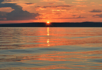 The sun, which has almost set behind the high hills, turns the low clouds above the large lake orange.