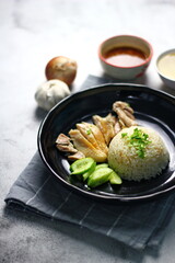 Hainanese chicken rice served on a black plate with dipping sauce and hot broth on white table.