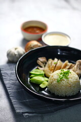 Hainanese chicken rice served on a black plate with dipping sauce and hot broth on white table.