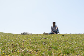 Fototapeta na wymiar Guy sitting far looking straight,left arm on knee in green grassland under blue sky in high altitude region in sunny day