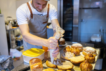 A male baker in a small artisan bakery fills brioches with cream. Front view.